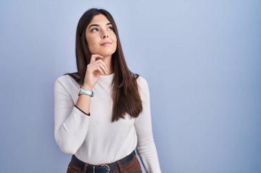 Mavi arka planda duran genç esmer kadın, elleri çenesinde soru sorarken, düşünceli bir yüz ifadesi vardı. Düşünceli bir yüzle gülümsüyor. şüphe kavramı. 