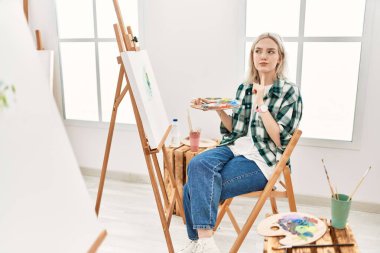 Young artist woman painting on canvas at art studio thinking concentrated about doubt with finger on chin and looking up wondering 