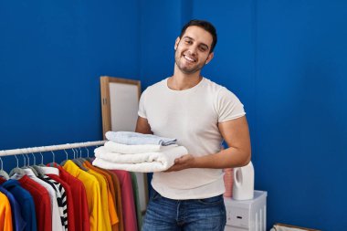 Sakallı, çamaşırhanede katlanmış temiz havluları tutan genç İspanyol adam yüzünde mutlu ve havalı bir gülümsemeyle gülümsüyor. diş gösterme. 