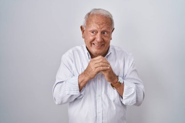 Senior man with grey hair standing over isolated background laughing nervous and excited with hands on chin looking to the side 