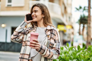 Elinde kahve tutan genç sarışın kadın caddede akıllı telefondan konuşuyor.