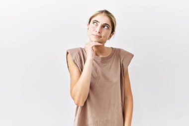 Soyutlanmış arka planda, eli çenesinde soru soran, düşünceli bir genç sarışın kadın. Düşünceli bir yüzle gülümsüyor. şüphe kavramı. 