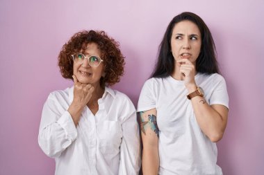 İspanyol anne ve kızı pembe arka plan üzerine günlük beyaz t-shirt giyiyorlar. Bir soru hakkında endişeleniyorlar. Endişeli ve sinirliler. 