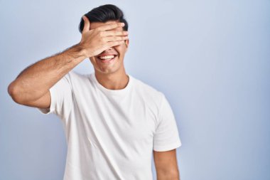 Mavi arka planda dikilen İspanyol bir adam. Gülümsüyor ve gülümsüyor. Gözlerini şaşkınlıkla kapatıyor. kör kavram. 