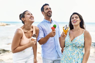 Üç genç İspanyol arkadaş gülümseyerek kumsalda dondurma yiyor..