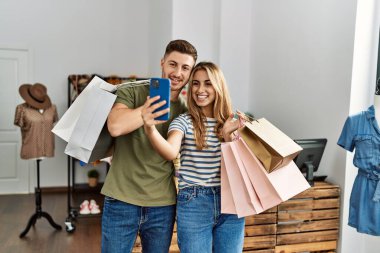 Young hispanic customer couple holding shopping bags and make selfie by the smartphone at clothing store.