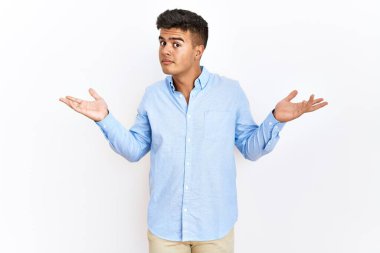 İzole edilmiş arka plan üzerinde duran İspanyol gömlekli genç adam elleri ve kolları havada, kafası karışık bir ifade takındı. şüphe kavramı. 