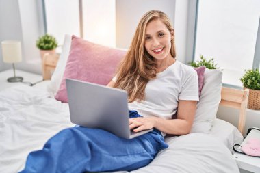 Yatak odasında dizüstü bilgisayar kullanan genç sarışın kadın.