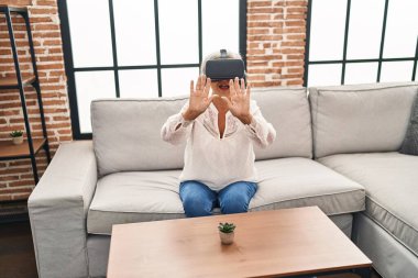Evde sanal gerçeklik gözlüğü kullanan orta yaşlı bir kadın video oyunu oynuyor.