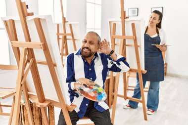 Senior artist man at art studio smiling with hand over ear listening an hearing to rumor or gossip. deafness concept. 
