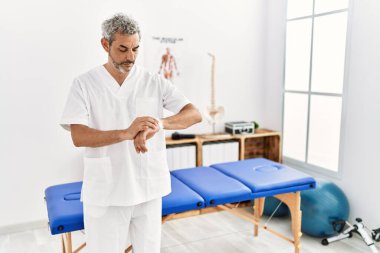 Orta yaşlı İspanyol terapist. Ağrı kesici kliniğinde çalışıyor. Kol saatini kontrol ediyor. 