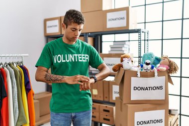 Bağışlarda gönüllü tişört giyen yakışıklı İspanyol genç adam kol saatinde saati kontrol ediyor, rahat ve kendinden emin. 