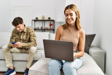 Genç İspanyol çift dizüstü bilgisayar ve akıllı telefon kullanarak mutlu bir şekilde gülümsüyor. Evdeki kanepede oturuyor..