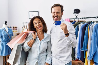 Hispanic middle age couple holding shopping bags and credit card sticking tongue out happy with funny expression. emotion concept. 