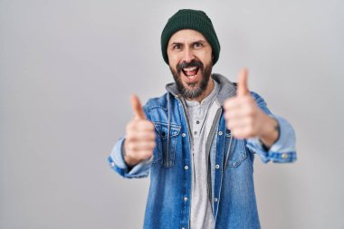 Young hispanic man with tattoos wearing wool cap approving doing positive gesture with hand, thumbs up smiling and happy for success. winner gesture. 