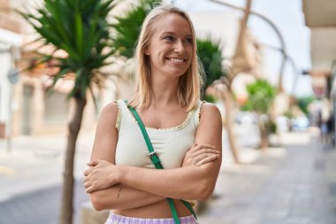 Genç sarışın kadın gülümsüyor. Kollarını kavuşturup caddede duruyor.