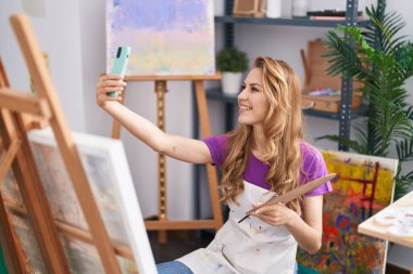 Genç sarışın sanatçı gülümsüyor. Sanat stüdyosundaki akıllı telefondan özçekim yapıyor.