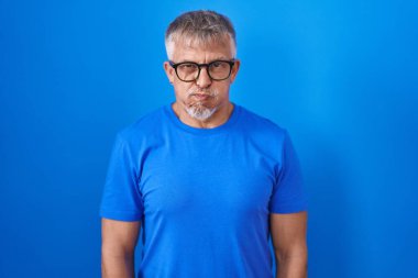 Mavi arka planda dikilen gri saçlı, komik yüzlü İspanyol bir adam. Havayla şişmiş ağız, çılgın bir ifade.. 