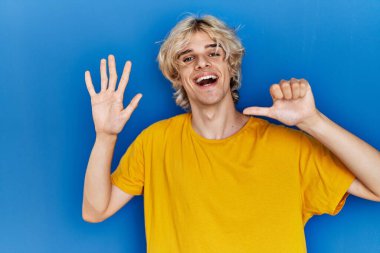 Genç modern bir adam mavi arka planda duruyor. Altıncı parmaklarını gösterip işaret ederken kendinden emin ve mutlu bir şekilde gülümsüyor.. 