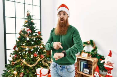 Uzun sakallı kızıl saçlı adam Noel ağacının yanında Noel şapkasını takıyor. Aceleyle saati, sabırsızlığı, gecikmeyi izlemek için işaret ediyor. 