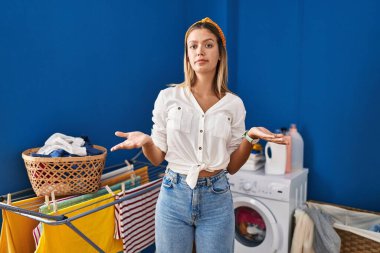 Young blonde woman at laundry room clueless and confused with open arms, no idea concept. 