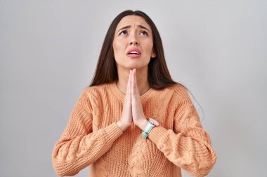 Genç esmer kadın beyaz arka planda duruyor. Elleriyle yalvarıyor ve dua ediyor. Yüzünde umut ifadesi var. Yalvarıyor. 