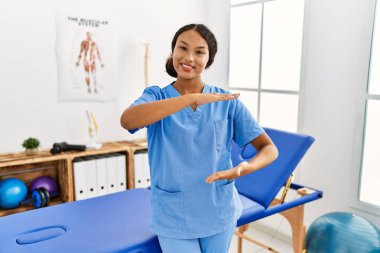 Güzel İspanyol fizyoterapist kadın. Ağrı kesici kliniğinde. Elleriyle büyük ve büyük bir işaret, ölçü sembolü gösteriyor. Kameraya bakıp gülümsüyor. ölçüm kavramı. 