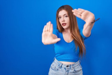 Mavi arka planda duran kızıl saçlı kadın el ve parmaklarıyla çerçeve yapıyor, kamera bakış açısı var. 