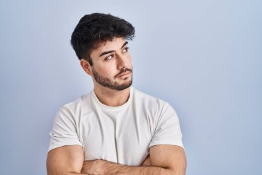 Hispanic man with beard standing over white background looking to the side with arms crossed convinced and confident 
