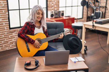 Müzik stüdyosunda dizüstü bilgisayar kullanarak klasik gitar çalan gri saçlı kadın müzisyen.