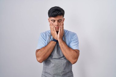 Beyaz arka planda önlük giyen İspanyol genç adam yorgun eller yüzünü, depresyonu ve üzüntüyü örten, sorun için üzgün ve tedirgin 