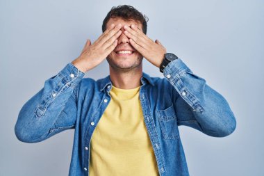 Mavi arka planda dikilen genç İspanyol adam, neşeli ve komik gülümseyen ellerle gözlerini kapatıyor. kör kavram. 