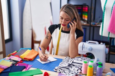 Young beautiful hispanic woman tailor talking on smartphone writing on notebook at clothing factory