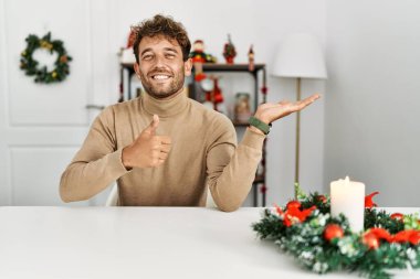 Yılbaşı süslemeleriyle masada oturan sakallı yakışıklı genç adam avuç içi gösteriyor ve baş parmaklarıyla iyi bir hareket yapıyor, mutlu ve neşeli gülümsüyor. 