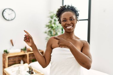 Genç Afro-Amerikan kadın havluyla güzellik merkezinde duruyor gülümsüyor ve kameraya bakıyor iki eliyle ve parmaklarıyla yandan işaret ediyor.. 