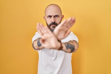 Young hispanic man with tattoos standing over yellow background rejection expression crossing arms and palms doing negative sign, angry face 