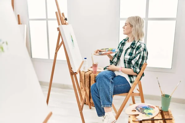 Young artist woman painting on canvas at art studio looking to side, relax profile pose with natural face and confident smile.