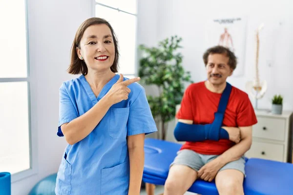 Middle age doctor woman with patient with arm injury at rehabilitation clinic smiling cheerful pointing with hand and finger up to the side