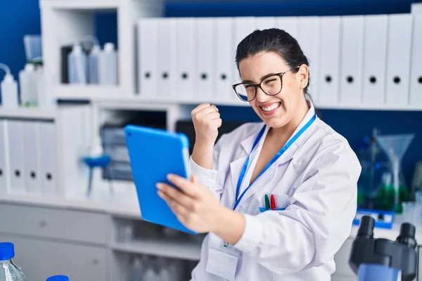 Young brunette woman working at scientist laboratory using tablet screaming proud, celebrating victory and success very excited with raised arm