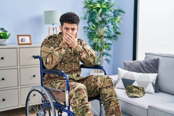 Arab man wearing camouflage army uniform sitting on wheelchair shocked covering mouth with hands for mistake. secret concept.