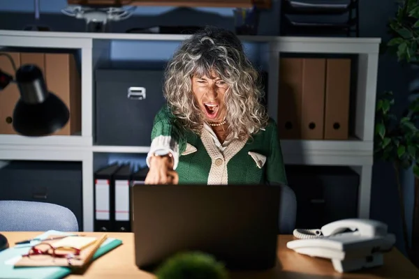 Middle age woman working at night using computer laptop pointing displeased and frustrated to the camera, angry and furious with you