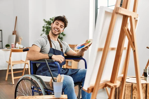 Young hispanic man sitting on wheelchair painting at art studio winking looking at the camera with sexy expression, cheerful and happy face.