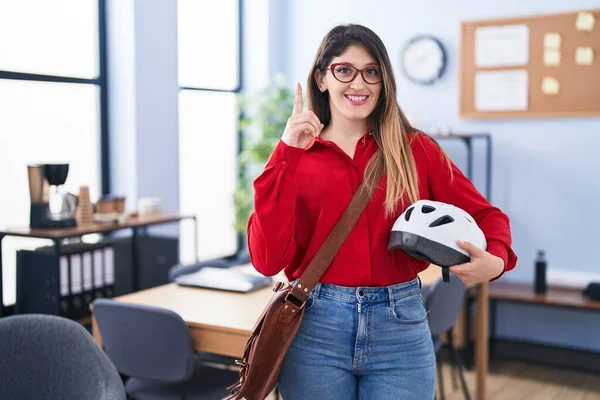 Young brunette woman working at the office holding bike helmet surprised with an idea or question pointing finger with happy face, number one