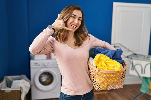 Young woman holding laundry basket smiling doing phone gesture with hand and fingers like talking on the telephone. communicating concepts.