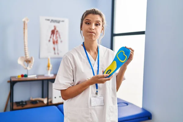 Young woman holding shoe insole at physiotherapy clinic puffing cheeks with funny face. mouth inflated with air, catching air.