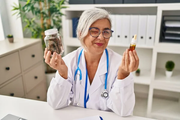 Middle age woman doctor holding cbd oil winking looking at the camera with sexy expression, cheerful and happy face.