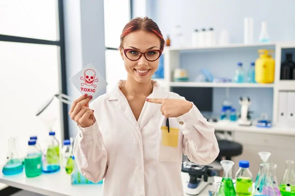 Young caucasian woman working at scientist laboratory holding toxic label pointing finger to one self smiling happy and proud