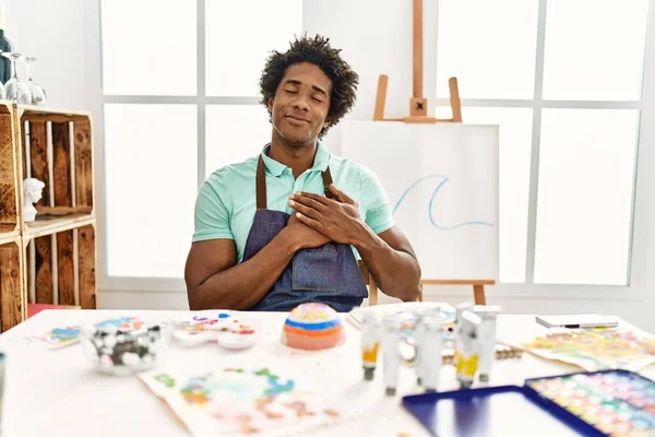 年轻的非洲裔美国人坐在艺术工作室的桌子上微笑着 双手放在胸前 闭上了眼睛 脸上挂着感激的手势 健康概念 — 图库照片