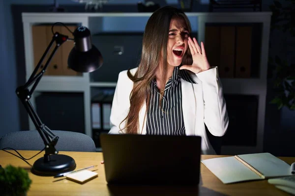 Young brunette woman working at the office at night with laptop shouting and screaming loud to side with hand on mouth. communication concept.