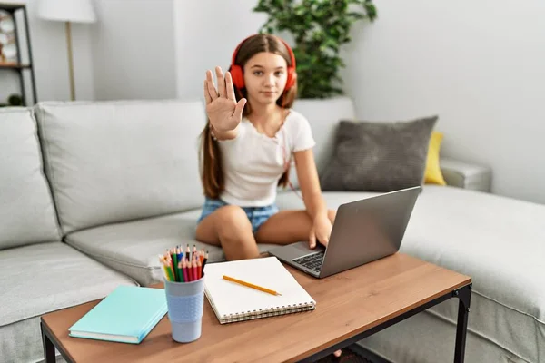 Young brunette teenager doing homework with laptop at home with open hand doing stop sign with serious and confident expression, defense gesture